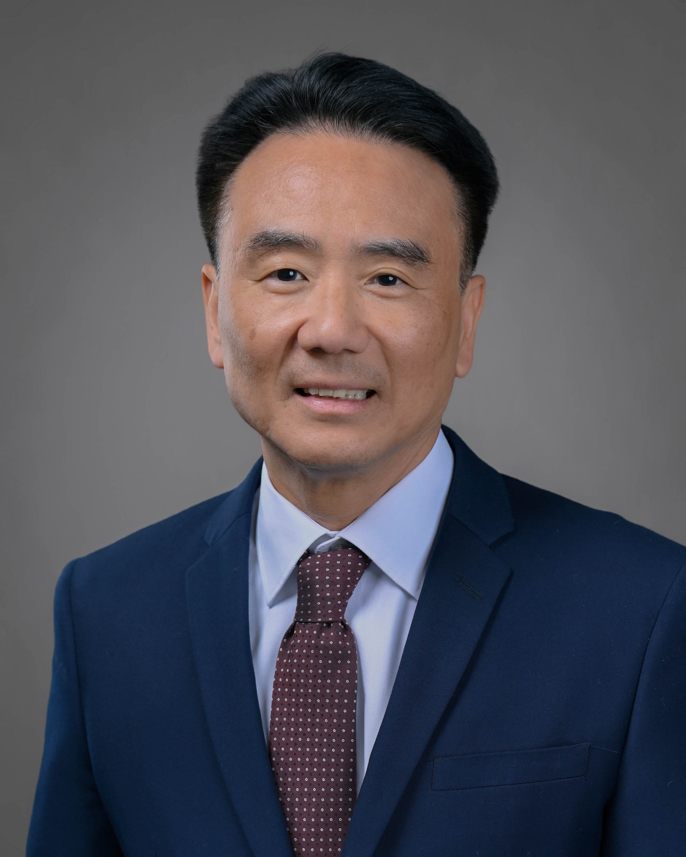 Kind looking man with dark hair in a dark blue suit with light blue shirt and dark red tie.